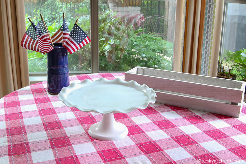 white scalloped cake stand, rectangle wood box, and blue vase with American flags on red check tablecloth 