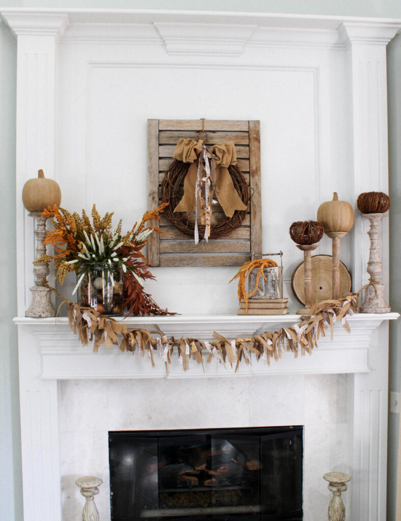 large glass jar filled with faux foliage and paper pumpkin on wood candlestick on mantel with grapevine wreath and burlap garland