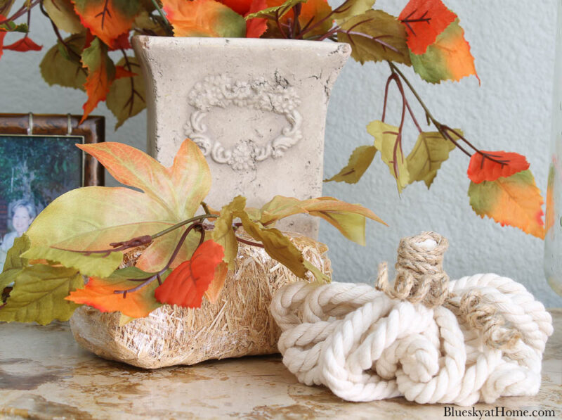 white rope pumpkin, small hay bale, adn stone vase with orange faux florals.