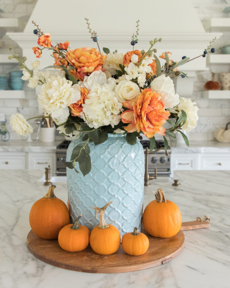 light blue vase with white and orange flowers and pumpkins