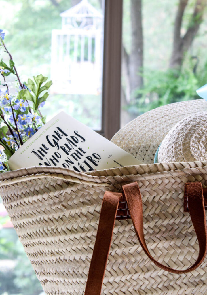 French woven tote basket bag with leather handles, straw hat, blue shawl, and book
