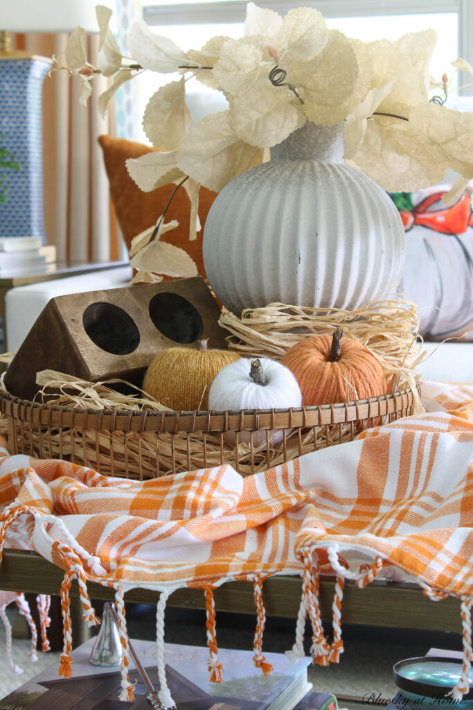 round basket with raffia in front of white sofa with white vase and cream faux leaves and sugar mold and yarn pumpkins