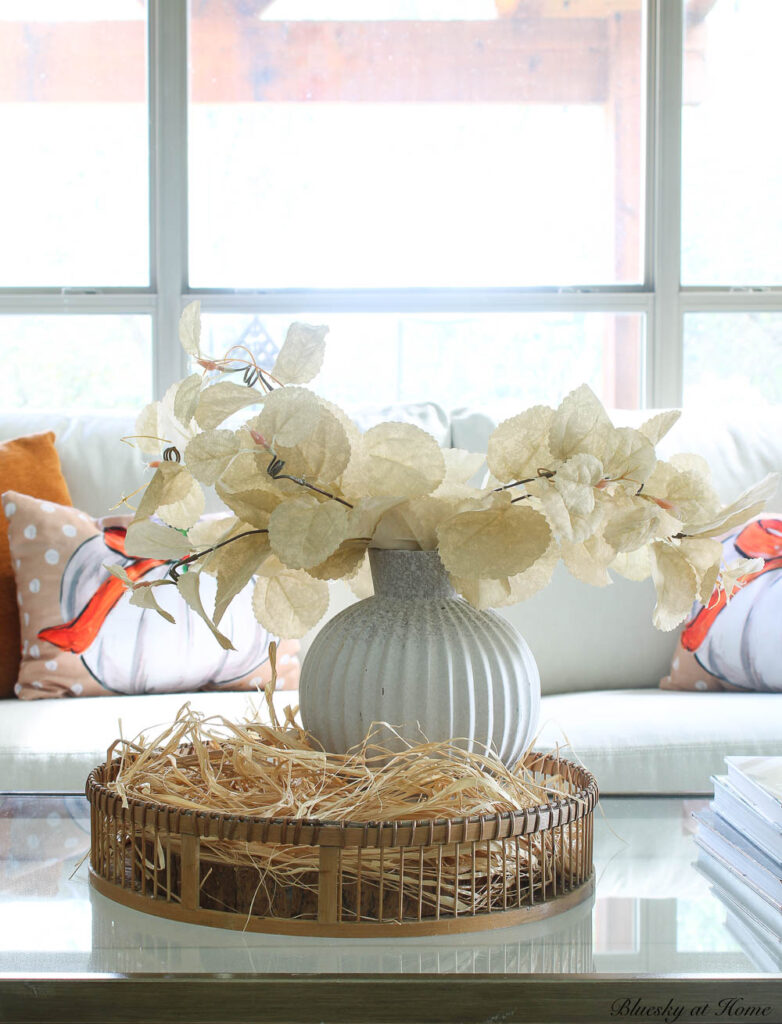 round basket with raffia in front of white sofa with white vase and cream faux leaves