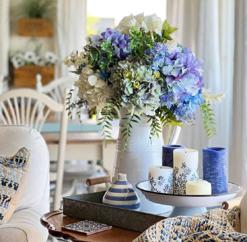 arrangement of blue hydrangeas in a white pitcher with blue candles and blue and white stripe vase in a metal tray