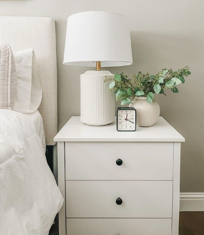 white modern lamp on a white nightstand with a vase of greenery and a small clock