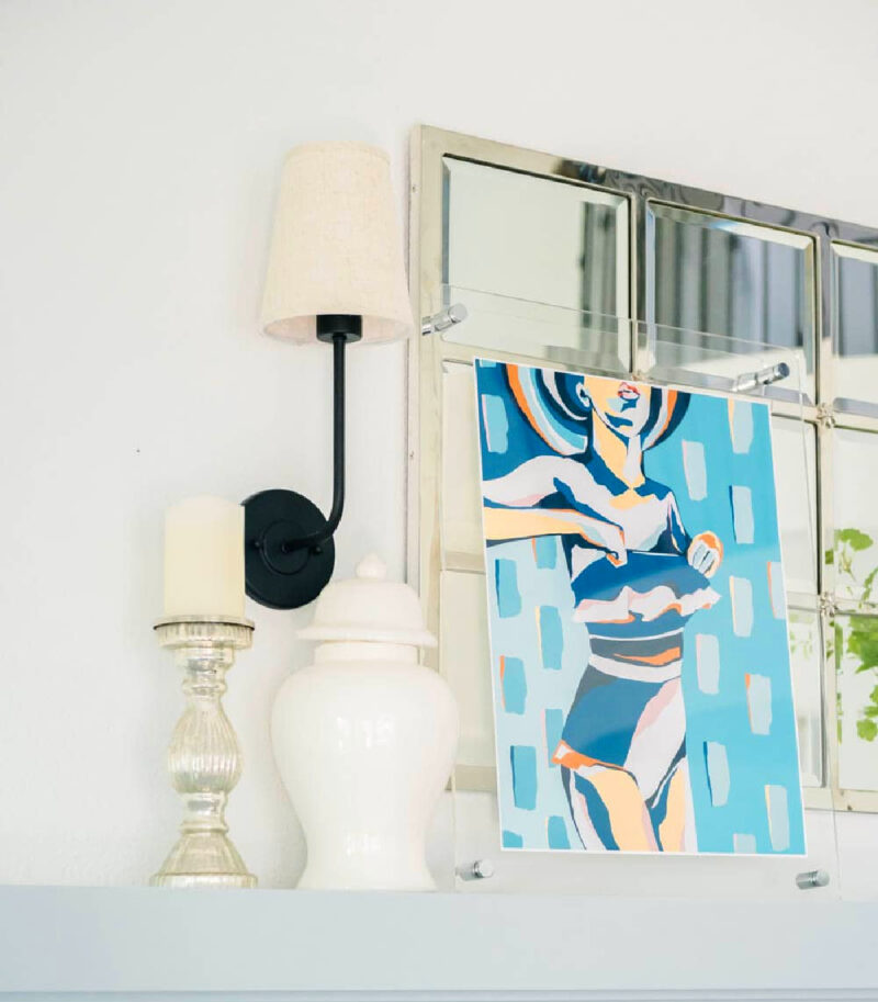 mantel with a white lamp, wood candleholder, and artwork of a women in a blue bathing suit