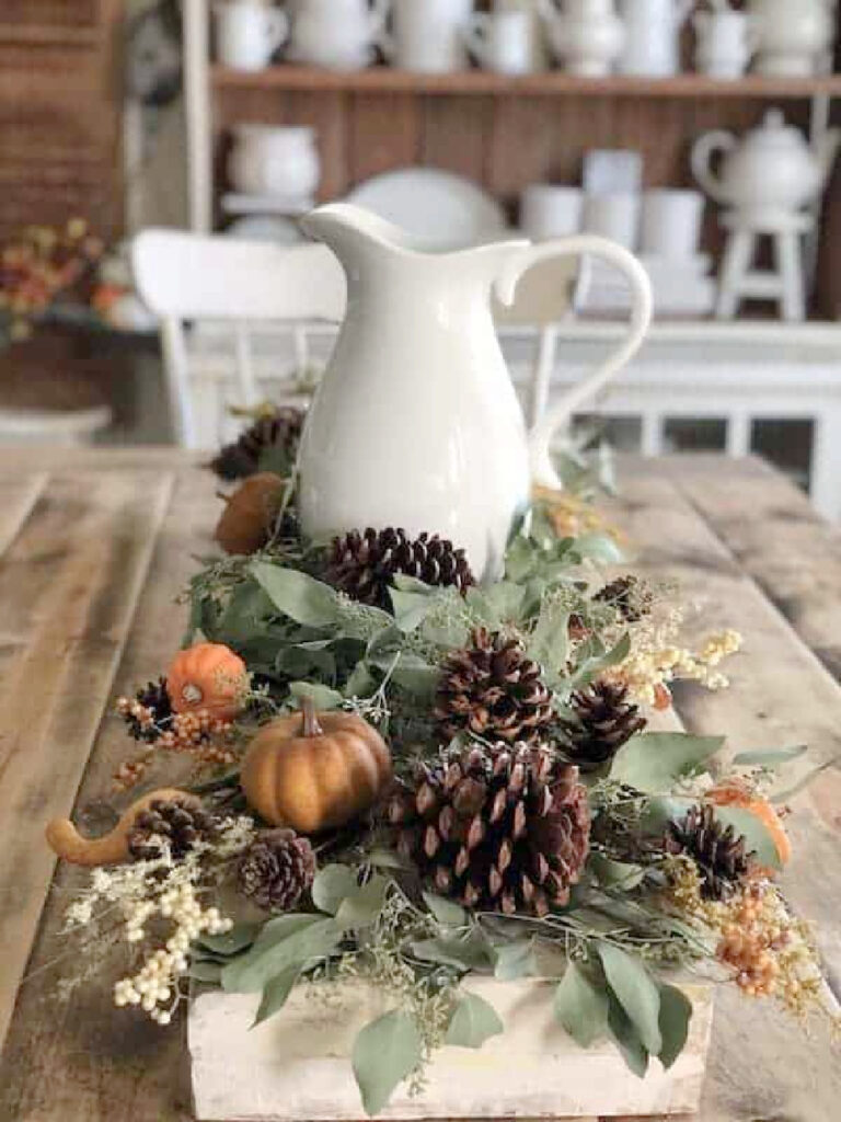 white pitcher on wood table with greenery, pinecones, and pumpkins