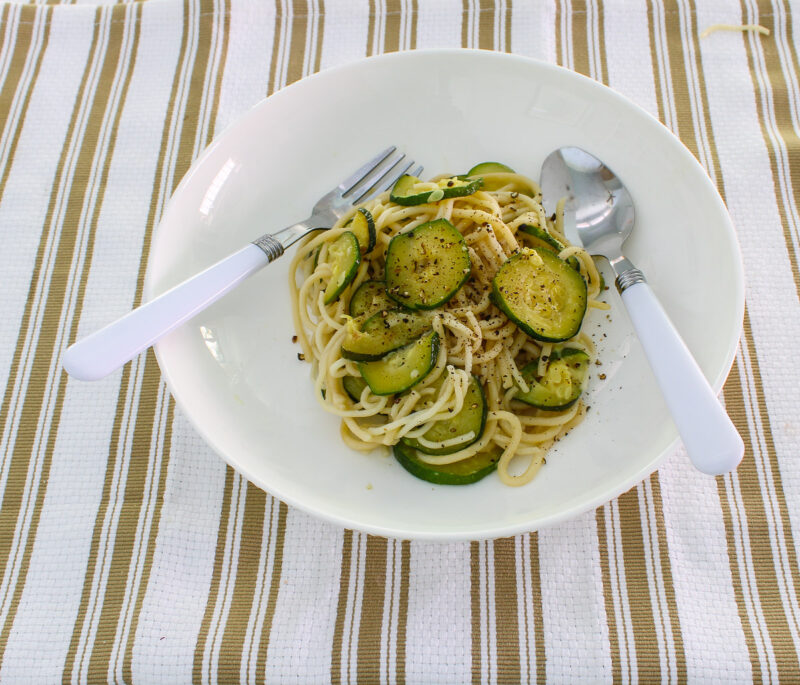 white bowl of spaghetti with sliced cooked zucchini and a fork and spoon with white handles