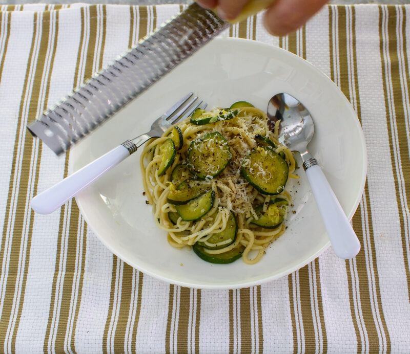 grating cheese on top of white bowl of spaghetti with sliced cooked zucchini and a fork and spoon with white handles