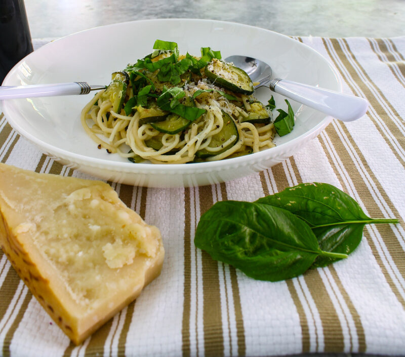 white bowl of spaghetti with sliced cooked zucchini and a fork and spoon with white handles with a chunk of parmesan cheese and basil leaves