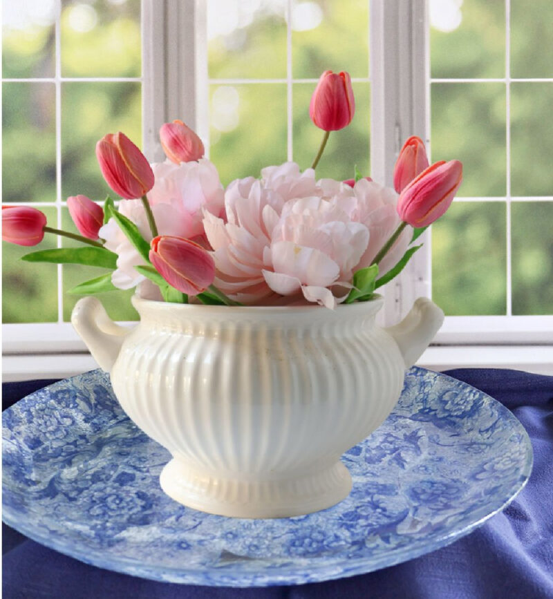 pink flowers in a white tureen on a blue decoupage platter in front of a window