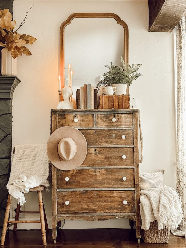 rustic scene with chest of drawers, mirror, chair, stool, books, blankets, plant