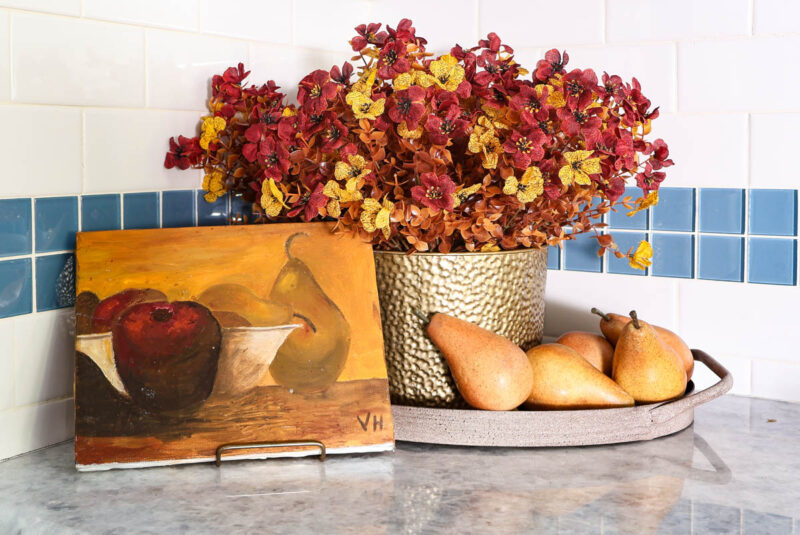 faux flower arrangement in brass container on tray with wood pears and still life painting